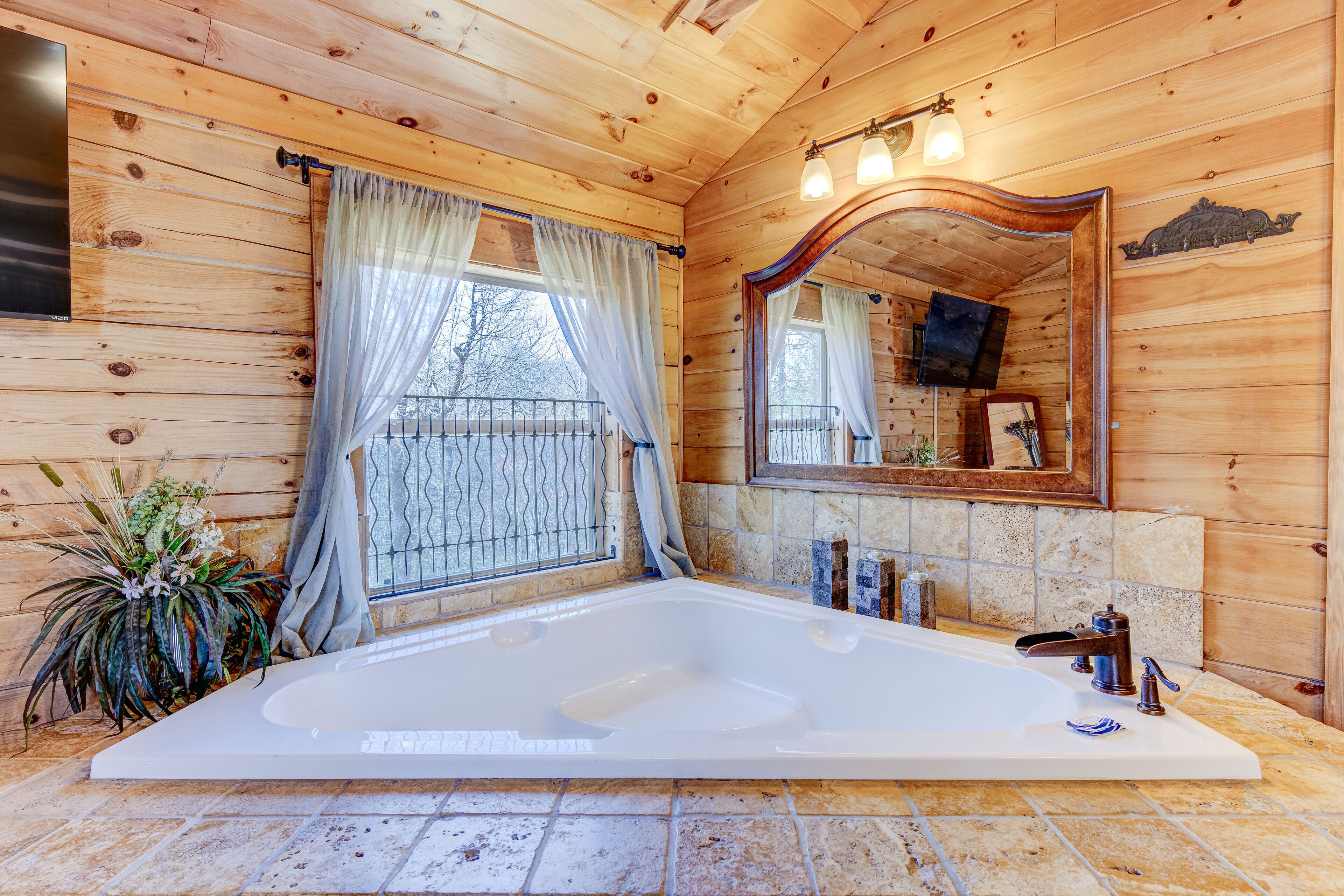 A wooden cabin bathroom with a large bathtub, a mirror, and a window with curtains.