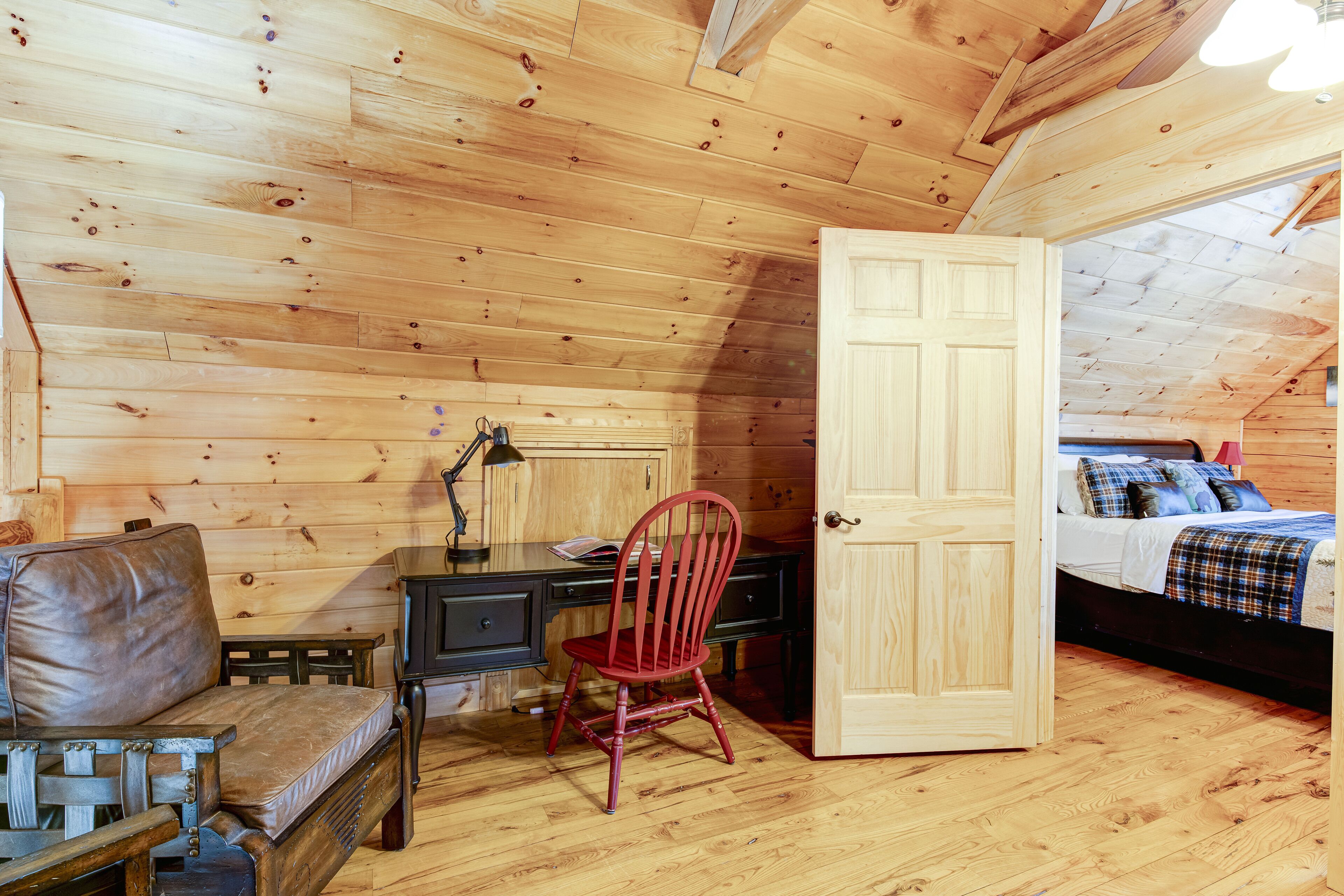 Ein gemütliches Zimmer mit Holzbalkendecke, einem Schreibtisch mit Stuhl, einem Bett mit kariertem Überwurf und einer Tür zu einem weiteren Raum.