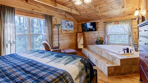 Ein Holzblockhaus-Interieur mit einem großen Bett, einem Fernseher, einem Badezimmer mit Badewanne und einem Fenster mit Vorhängen.