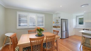A modern kitchen with white cabinets, a stainless steel refrigerator, and a dining area with wooden chairs and a table. - Sunny Time - Avoca Beach (Avoca)