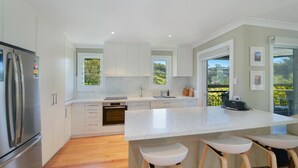 A modern kitchen with white cabinets, a large island with bar stools, a built-in oven, and a refrigerator. - Sunny Time - Avoca Beach (Avoca)