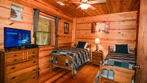 A cabin-style bedroom with two beds, a wooden dresser, a television, and a ceiling fan.