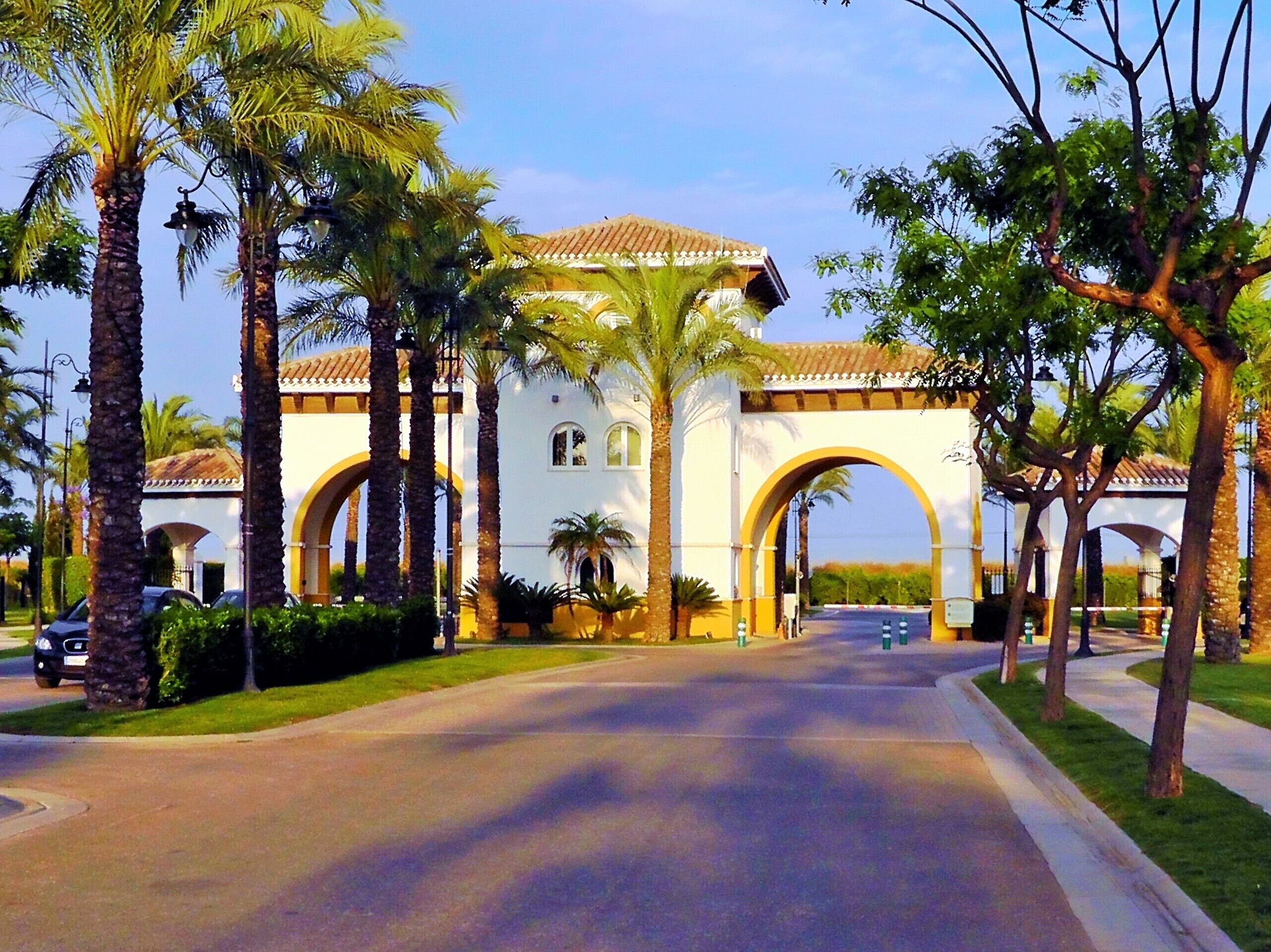 A white building with arched entrances, surrounded by palm trees and a paved road.