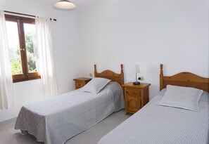 A room with two single beds, wooden headboards, bedside tables with lamps, a window with curtains, and a tiled floor.