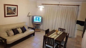 Una sala de estar con sofá, televisor, mesa de comedor con sillas y ventana con cortinas.