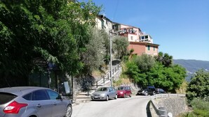 Uma estrada estreita com carros estacionados de um lado, um muro de pedra e um edifício com varandas ao fundo.