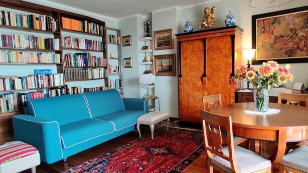 Ein gemütliches Wohnzimmer mit einem blauen Sofa, einem Holzesstisch mit Stühlen, einem Bücherregal voller Bücher und einem roten Teppich mit Muster.