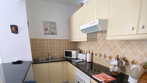 A compact kitchen with a microwave, toaster, and stove. There are two blue chairs and a small dining table. The kitchen has beige cabinets and a tiled backsplash.