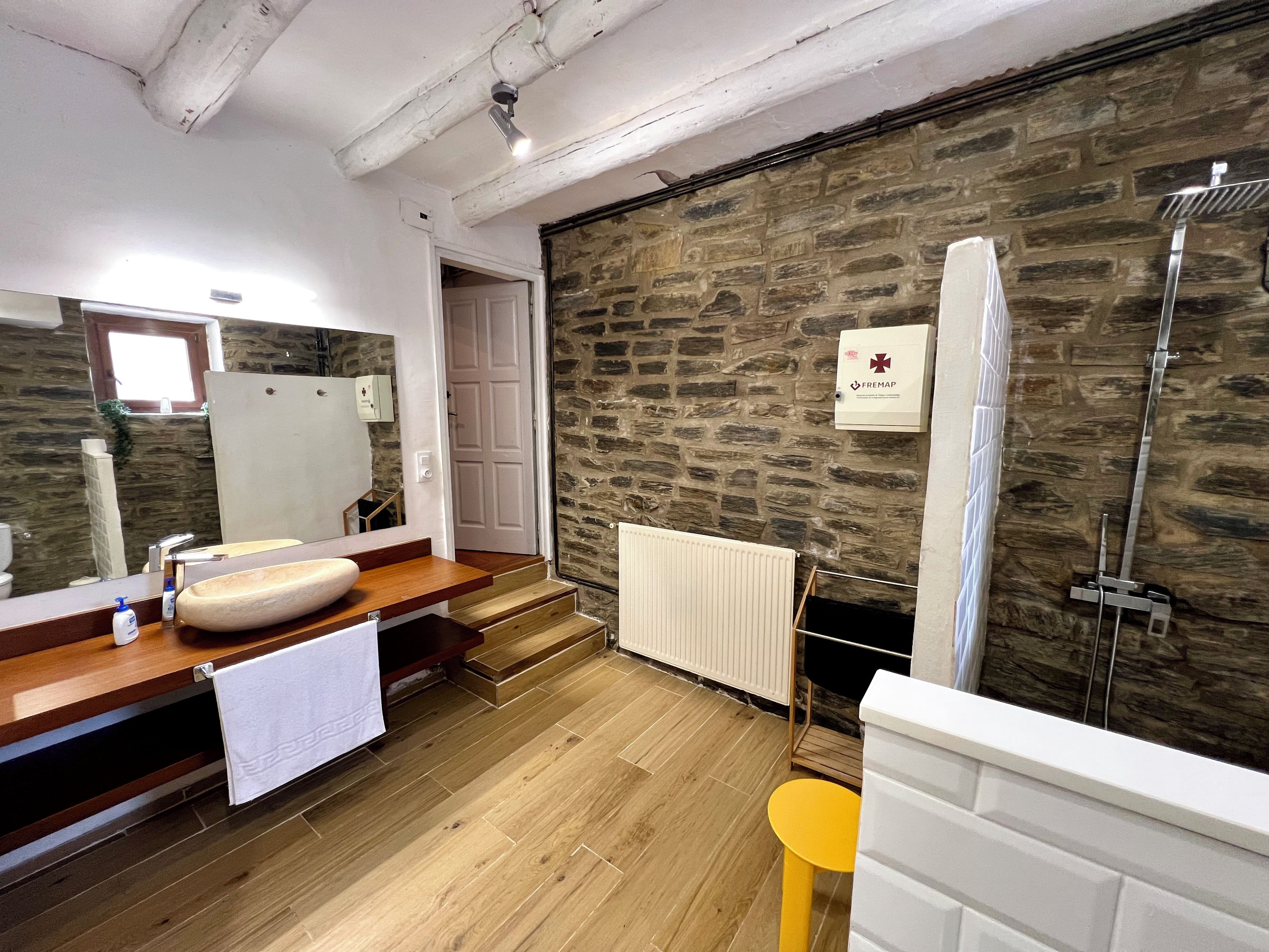 A modern bathroom with a stone wall, wooden flooring, a large mirror, a sink, and a shower.