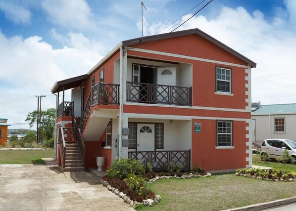 A two-story building with a balcony, a staircase, and a small garden area. - Breezy Apts (Four Roads)