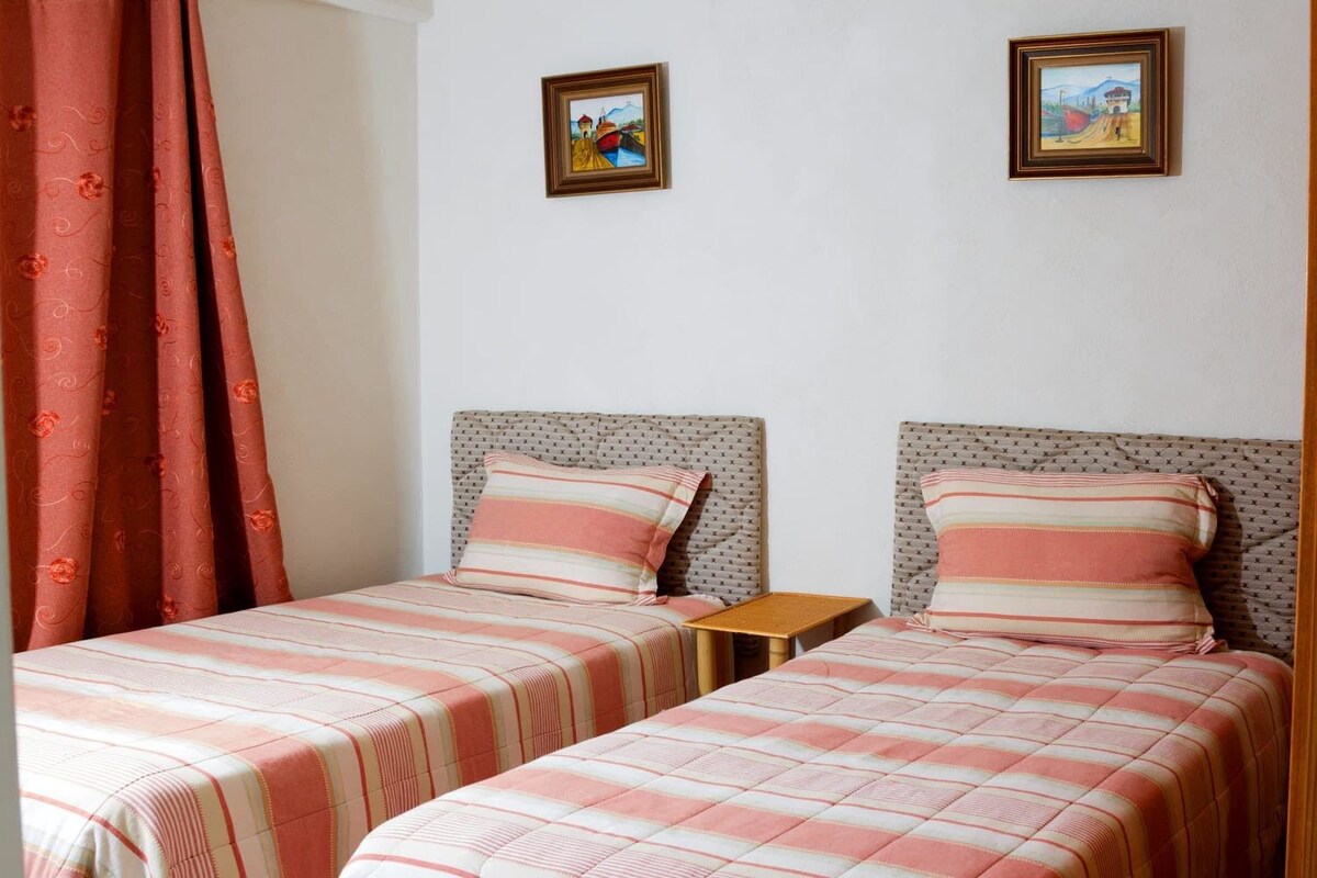 A room with two beds, each with striped bedding and matching pillows. There are two framed pictures on the wall, a small wooden table, and a red curtain.
