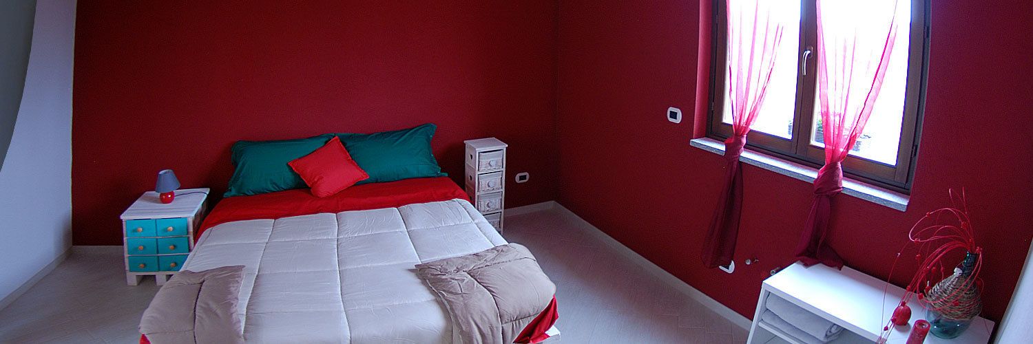 Un dormitorio con una pared roja, una cama con ropa de cama blanca y roja, una mesita de noche con lámpara y una ventana con cortinas.