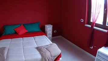 Un dormitorio con una pared roja, una cama con ropa de cama blanca y roja, una mesita de noche con lámpara y una ventana con cortinas.