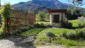 Una propiedad campestre con un portón de madera, un pequeño edificio de piedra con techo de tejas y montañas al fondo.