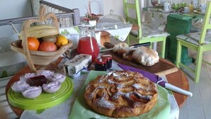 Una mesa de desayuno con una cesta de frutas, hogaza de pan, plato de cebollas rebanadas, frasco de mermelada y un pastel sobre una tabla de cortar verde.