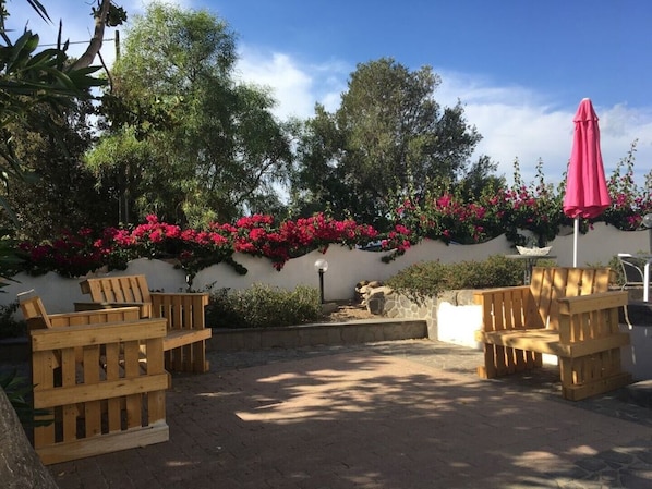 An outdoor patio area with wooden chairs and a pink umbrella, surrounded by lush greenery and flowering plants.