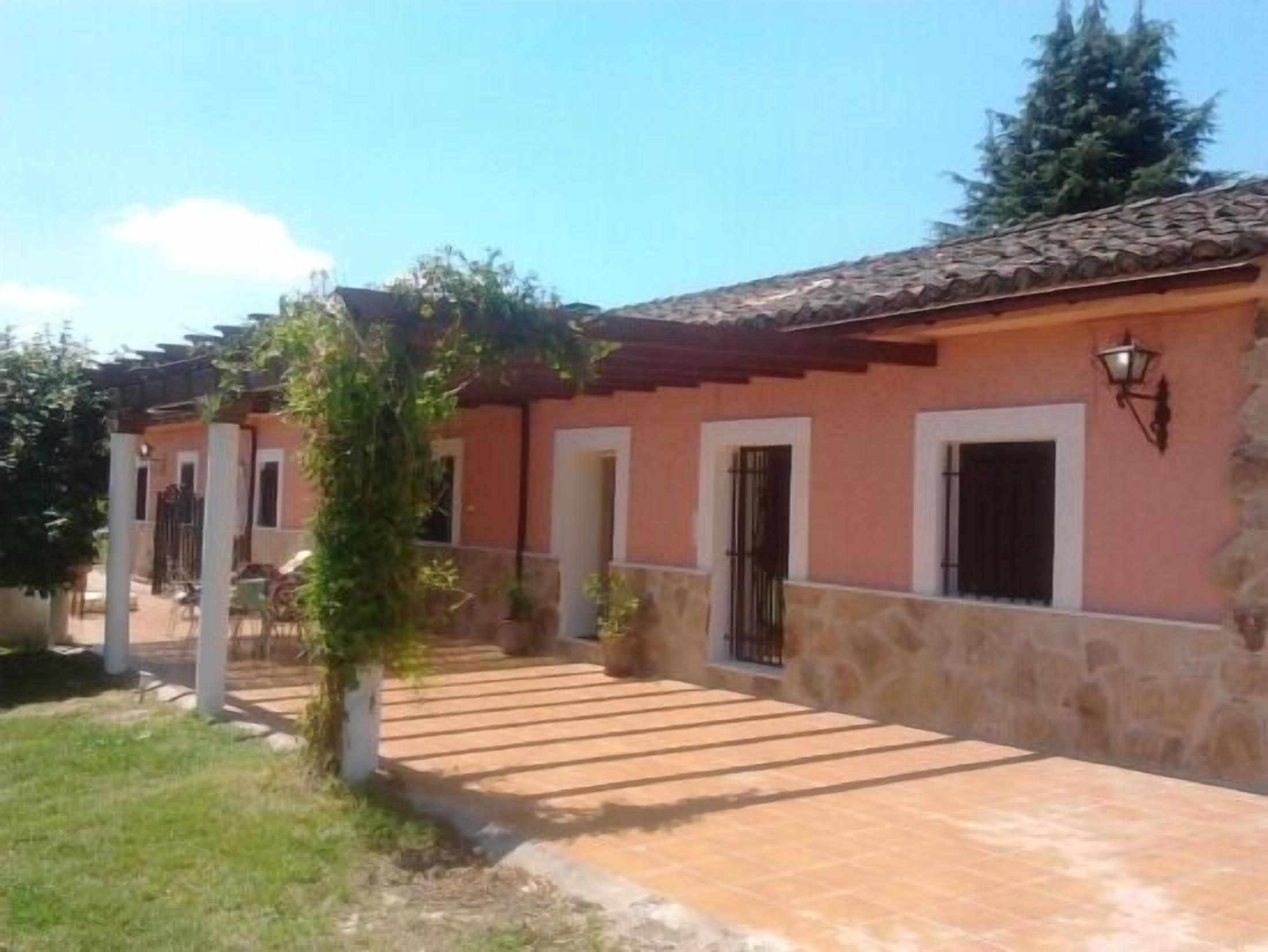 Un edificio con tejado de tejas, base de piedra y fachada rosa. Se observan dos puertas y un pequeño jardín con un árbol y un banco.