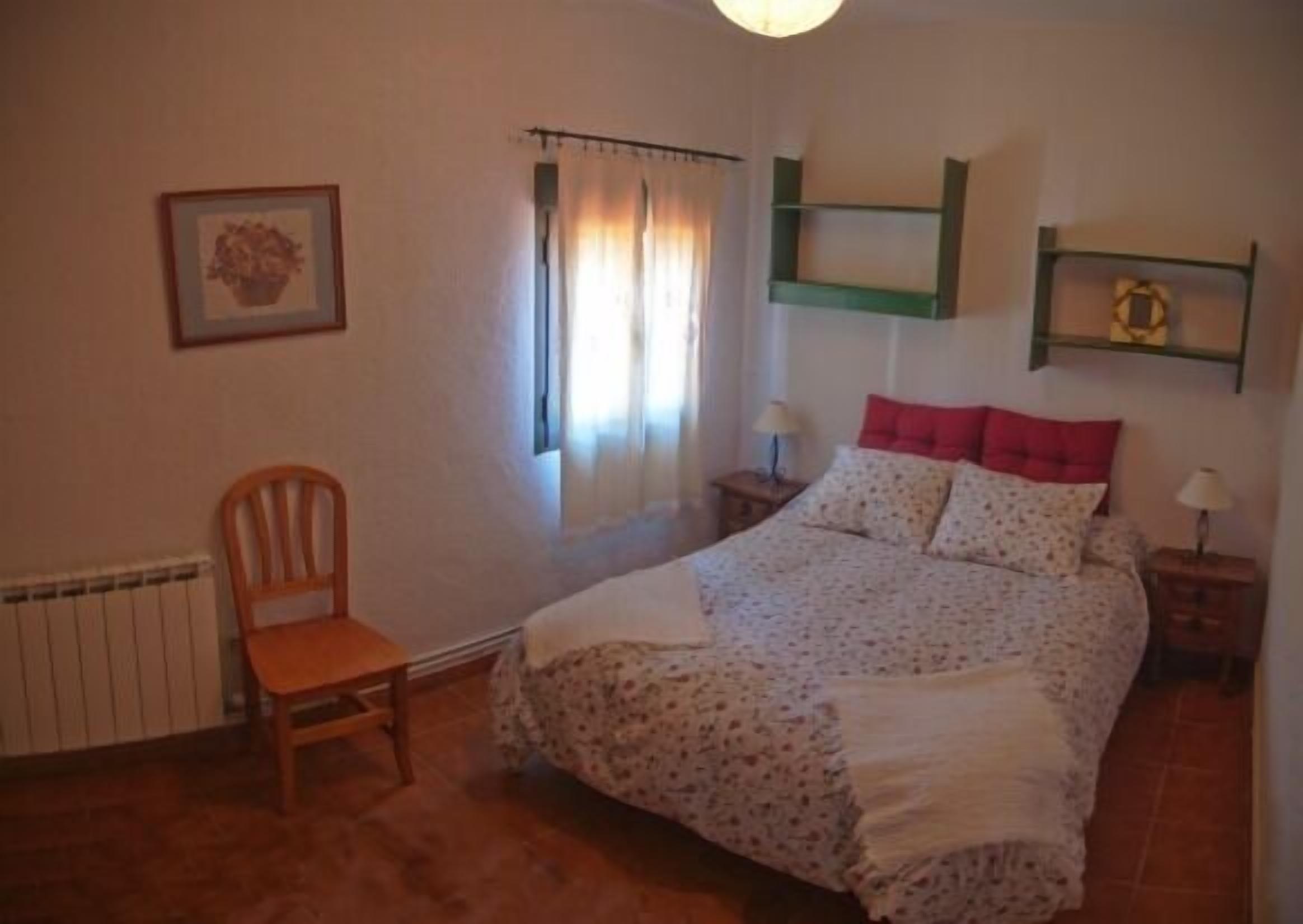 Un dormitorio con cama, una silla, un radiador, una ventana con cortinas y una estantería con un cuadro.