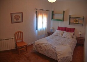 Une chambre à coucher avec un lit, une chaise, un radiateur, une fenêtre avec des rideaux et une étagère sur laquelle est posée une image.
