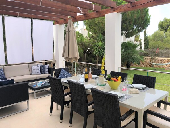 An outdoor patio with a dining table set for two, surrounded by black wicker chairs, a beige sofa with patterned cushions, and a wooden pergola overhead. - Villa Ohana · Stylish 4-Bed Retreat for 8 Guests with Pool in Sunny Cala d’Or (Cala d'Or)
