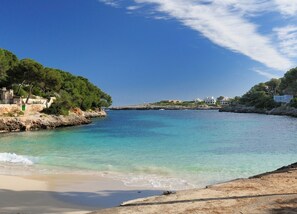 A coastal scene with clear turquoise water, sandy beach, and buildings along the shore. - Villa Ohana · Stylish 4-Bed Retreat for 8 Guests with Pool in Sunny Cala d’Or (Cala d'Or)