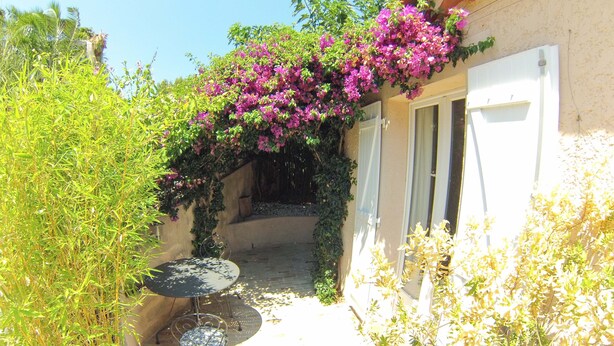 A sunlit courtyard with a table and chairs, surrounded by lush greenery and flowering plants. - Apartment in villa, sea view from bedrooms (Hyères)