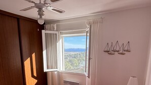A bedroom with a bed, a ceiling fan, a window with curtains, a radiator, and a wall-mounted shelf with decorative items. - Apartment in villa, sea view from bedrooms (Hyères)