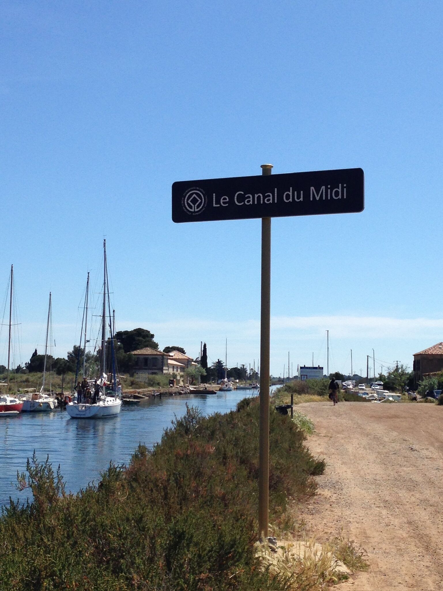 Une pancarte indiquant « Le Canal du Midi », avec des bateaux amarrés le long d’un canal, un chemin de terre et des bâtiments en arrière-plan.