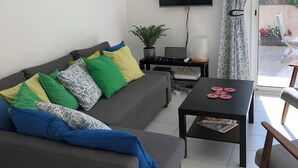 Un salon moderne avec un canapé gris, une table basse, un téléviseur et une vue sur l’extérieur par une fenêtre ornée de rideaux à motifs.