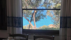 A dining table with four chairs, a window with a view of trees and a body of water, and a curtain with a blue pattern. - Villa "The lapwing one" (semi-detached portion of the sea) (Marina di Tertenia, Tertenia (OG))