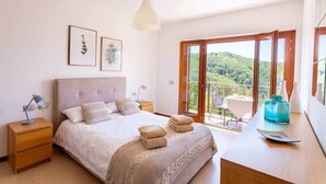 Een slaapkamer met een bed, nachtkastjes, een bureau met stoel, en een balkon met uitzicht op groen.
