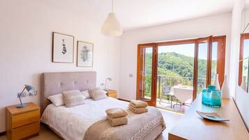 Une chambre à coucher comprenant un lit, des tables de chevet, un bureau avec une chaise et un balcon donnant sur un espace verdoyant.
