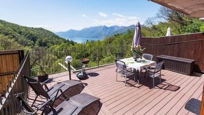 Een houten terras met een tafel en stoelen, met uitzicht op een bergachtig landschap en een waterpartij.