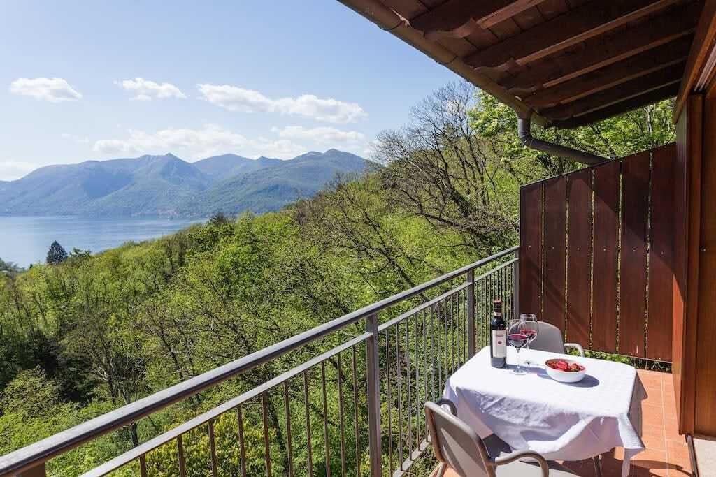 Un balcon avec une table dressée pour deux personnes, offrant une vue sur un paysage montagneux et un plan d’eau.