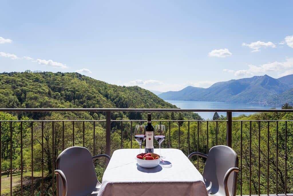 A table with a bottle and glasses, a bowl of fruit, and a scenic view of mountains and a lake.