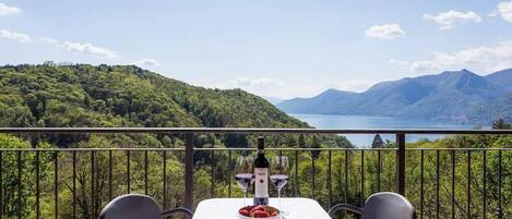 A table with a bottle and glasses, a bowl of fruit, and a scenic view of mountains and a lake.