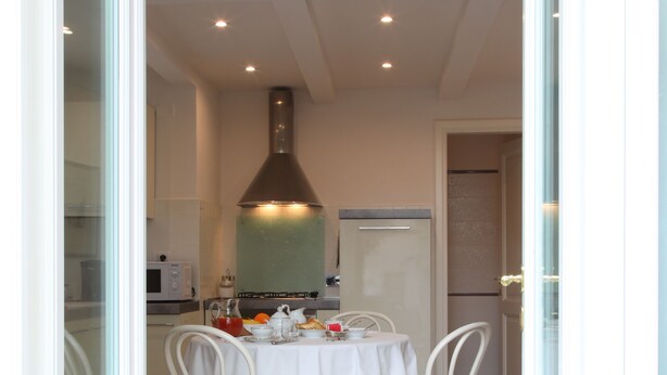 Zona pranzo con tavolo apparecchiato per quattro persone, sedie bianche, tovaglia bianca e in secondo piano una cucina con microonde e fornello.