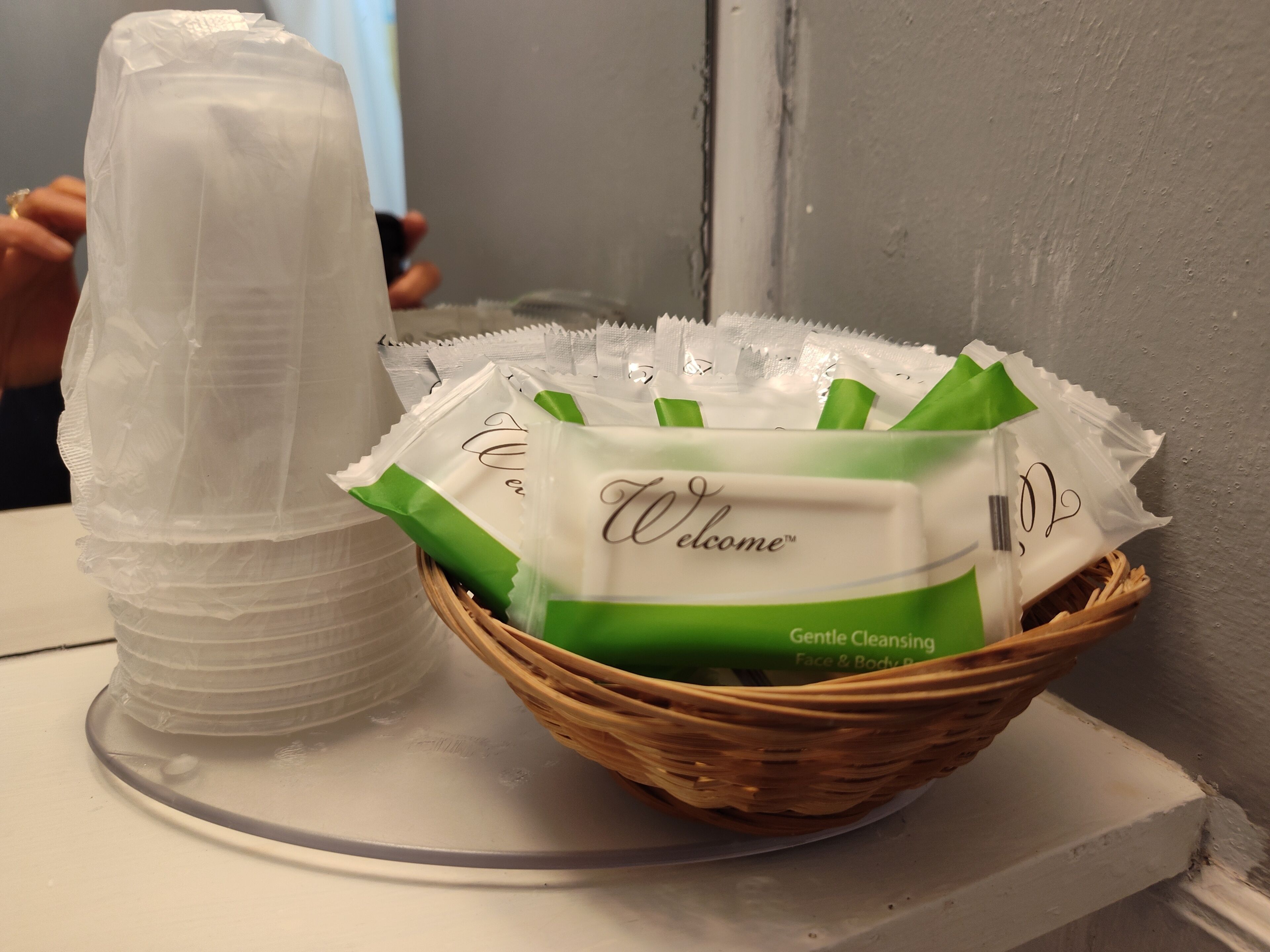 Una cesta con paquetes etiquetados 'Welcome Gentle Cleansing Face & Body' junto a una pila de vasos de plástico transparente.