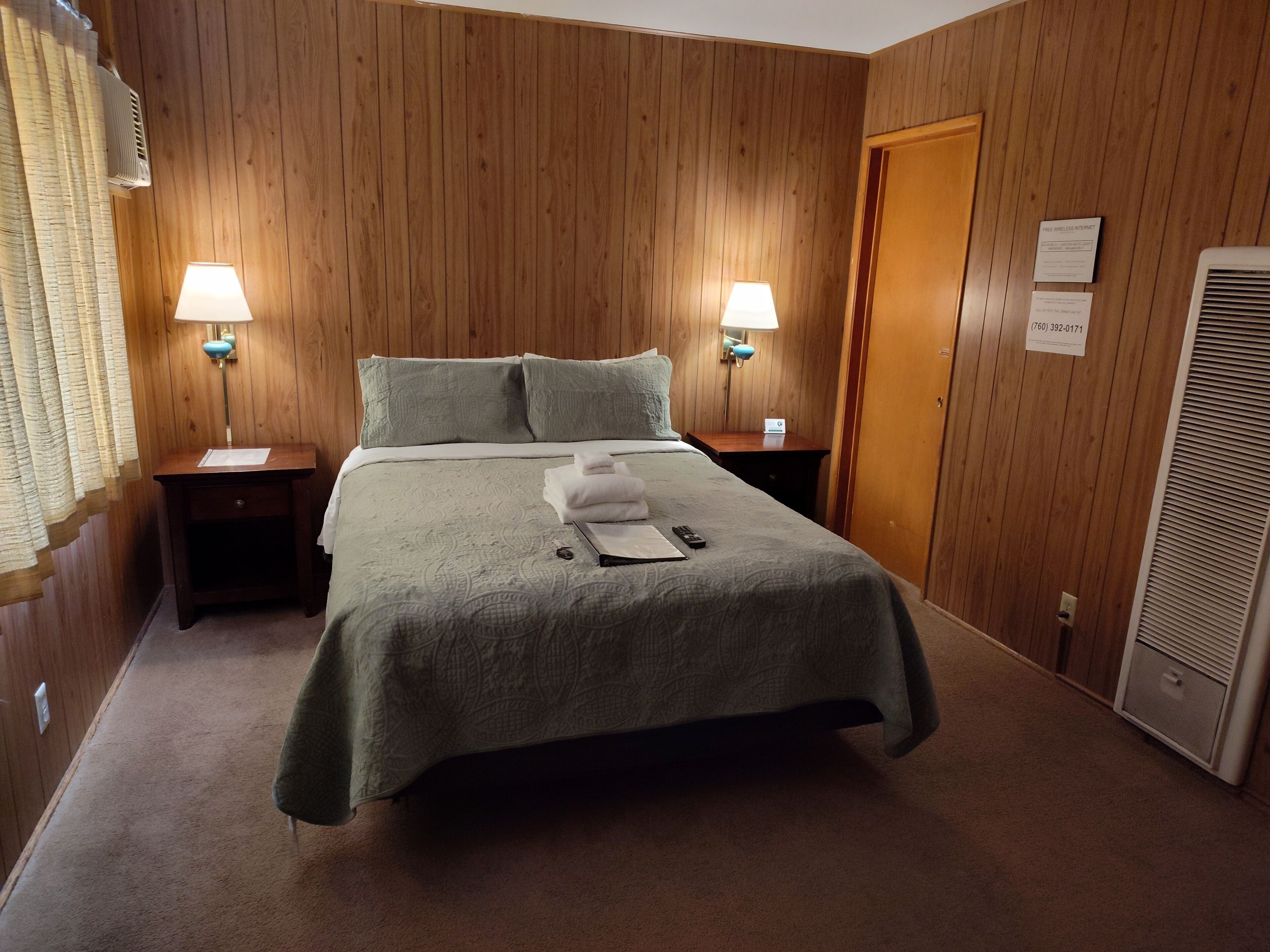 Een hotelkamer met een wand bekleed met houtpanelen, een bed met een groene dekbedovertrek, twee bedlampen, een nachtkastje en een deur met een bordje.