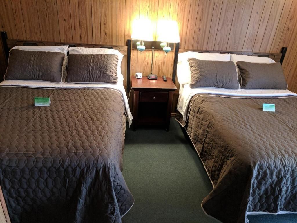 A room with two beds, each with a quilted bedspread and pillows, a nightstand with a lamp, and a small table with a radio and remote control.