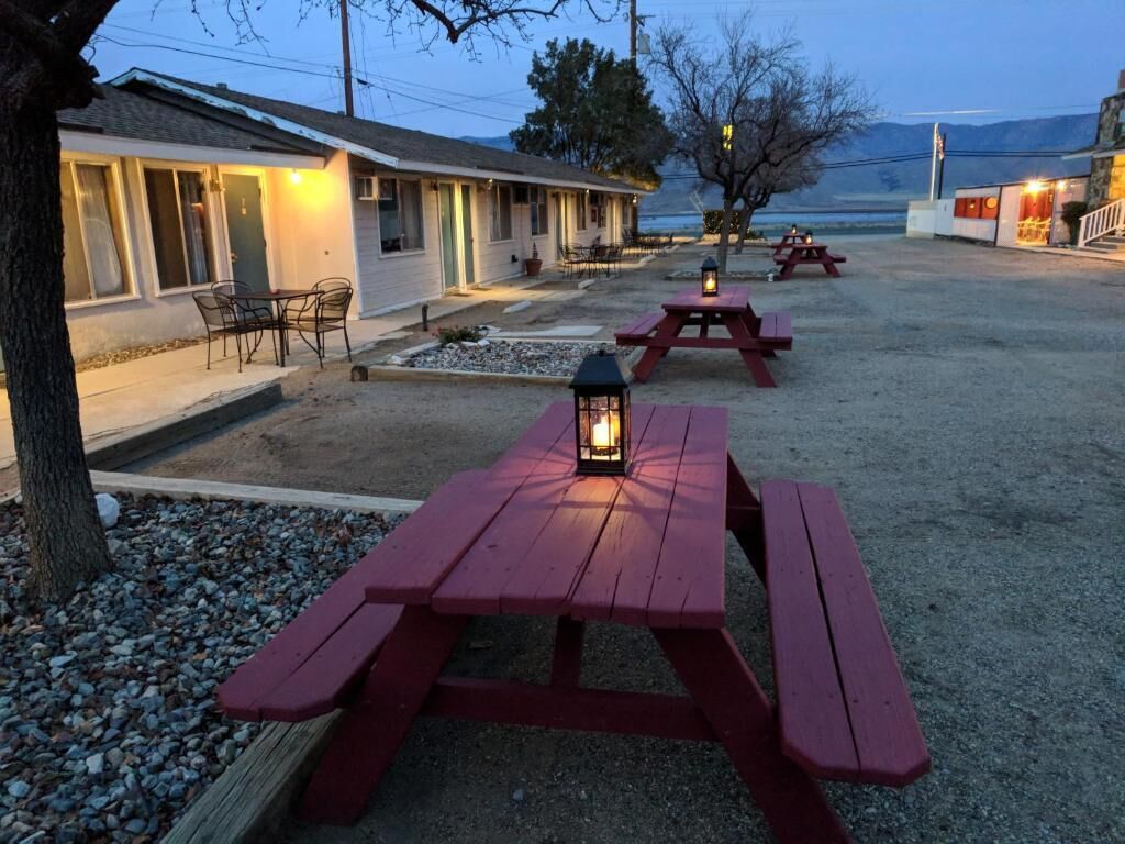 A rustic outdoor seating area with red picnic tables and lanterns, a building with a porch, and a view of the water in the background.