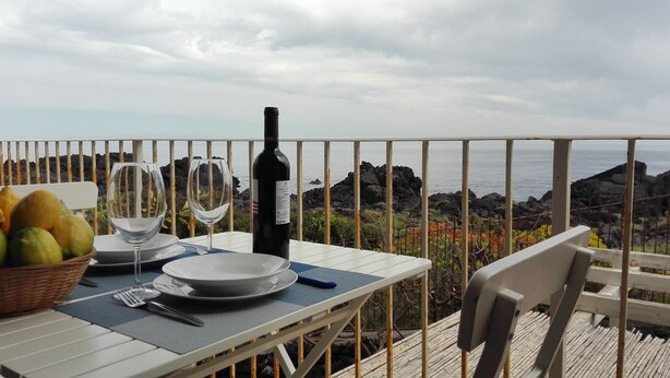 A table set for two with a bottle of wine, glasses, plates, and a basket of fruit on a balcony overlooking the sea. - Casa Pozzillo: two rooms with splendid sea right in front of the house! (Acireale)