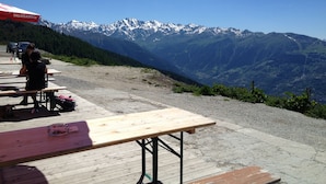 Un espace pique-nique avec des tables et des bancs en bois, un parasol rouge et une vue sur des montagnes enneigées.