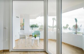 A balcony with a view of palm trees and buildings, a white chair, a small table with a plant, and a white sofa. - CRUISE APARTAMENT. First line beach. It will look like it's on a boat. (Málaga)