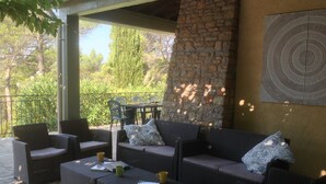 Eine überdachte Terrasse mit Korbmöbeln, einer Steinwand und Blick auf Bäume.