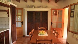 Uma sala de jantar com mesa e cadeiras de madeira, uma cesta de frutas e uma área de cozinha com armários e piso ladrilhado.