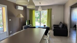 Un soggiorno moderno con tavolo da pranzo, divano e televisore. È presente un'ampia finestra con tende e un condizionatore d'aria a parete.
