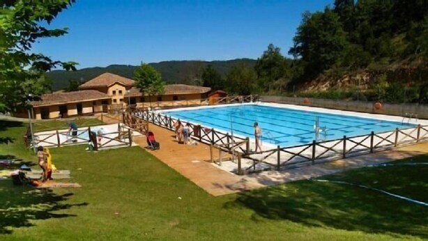Une piscine dotée d’une terrasse en bois et d’une zone délimitée par une clôture, entourée de pelouse et d’arbres.