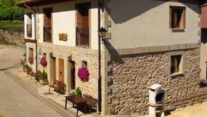 Un bâtiment à deux étages, doté d’une façade en pierre, d’un toit en tuiles et d’une cheminée. On y trouve un petit porche avec un banc et des plantes en pot.