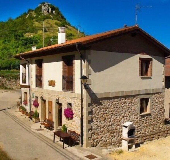 A two-story building with a stone facade, a tiled roof, and a chimney. There is a small porch with a bench and potted plants. - OFFER - Rural house (full rental) Gaztelubidea for 6 people (Bernedo)